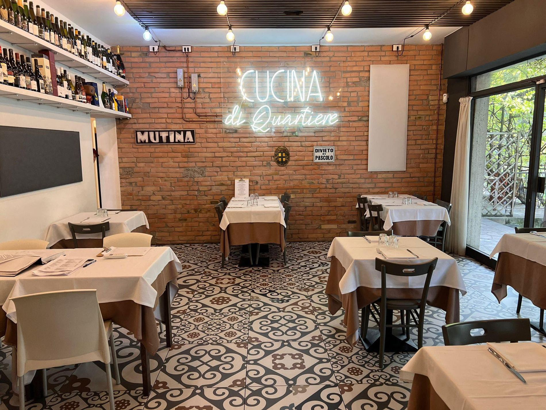 La sala della Cucina di Quartiere a Modena: parete di mattoni, insegna al neon, pavimento a cementine, tavoli apparecchiati con tovaglie bianche.